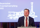 Verkehrsminister Christian Bernreiter steht an einem Rednerpult und hält eine Rede. Im Hintergrund eine Leinwand mit Aufdruck der Adler-Lok und Text „Bayerischer Eisenbahnempfang 2022“.