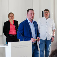 Ein Mann hält eine Rede. Im Hintergrund stehen eine Frau und ein weiterer Mann