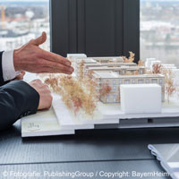 Auf einem Schreibtisch steht ein Modell eines Gebäudes. Zwei Hände zeigen darauf.